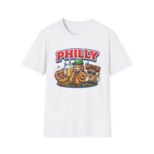 Op de afbeelding: Wit T-shirt met een cartoon illustratie. Het ontwerp toont het woord "PHILLY" in rood en blauw, een pretzel, een hotdog, een biertje en een personage met een zonnebril. De achtergrond toont een stadsgezicht.