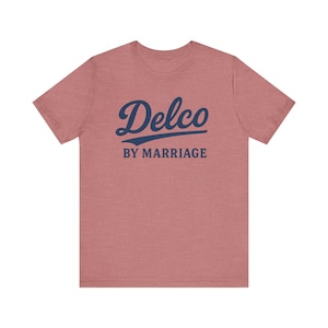 Peut inclure: Un t-shirt rose poussiéreux à manches courtes. Le texte bleu marine "Delco" est écrit en script, avec "BY MARRIAGE" en dessous en sans-serif. Le t-shirt est fait d'une matière douce.