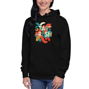 Könnte beinhalten: Schwarzer Kapuzenpullover mit dem Text "START FRESH" in farbenfrohen, pinselstrichartigen Buchstaben. Das Design weist Farbtöne von Orange, Türkis, Gelb und Rot auf. Der Hoodie hat eine Kapuze mit Kordelzug und eine Vordertasche.