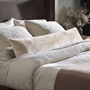 Peut inclure: Un long coussin traversin rectangulaire en velours crème placé horizontalement sur un lit. Le lit présente une housse de couette blanche texturée, des oreillers assortis et un jeté de lit crème uni et lisse au pied du lit.