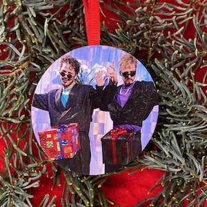May include: Round Christmas ornament featuring a photo of two people wearing sunglasses and holding wrapped gifts. The ornament has a red ribbon for hanging and is surrounded by evergreen branches on a red background.
