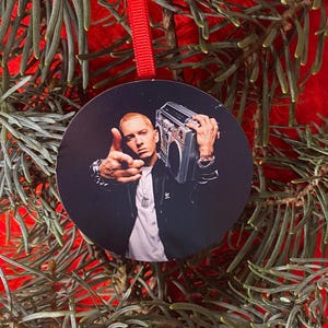 May include: Round Christmas ornament featuring a portrait of Eminem holding a boombox. The image is set against a black background. The ornament has a red ribbon for hanging and is surrounded by green pine needles.