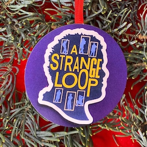 May include: A circular purple Christmas decoration featuring the text "A Strange Loop" in yellow. The ornament has a white outline in the shape of a head and includes small blue figures. A red ribbon is attached for hanging.