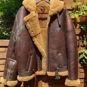 Pode incluir: Casaco de aviador de couro castanho com gola e forro de pele de carneiro. O casaco tem fecho de correr, bolsos e cintura com fivela. O casaco está pendurado num cabide de madeira contra uma cerca de madeira.