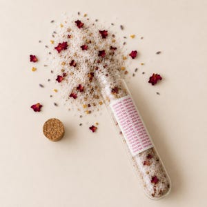 May include: A clear glass test tube filled with white bath salts, dried red rose petals, and small yellow flowers. A cork stopper sits nearby. A white label on the tube features the repeated pink text: I attract love, I attract love.