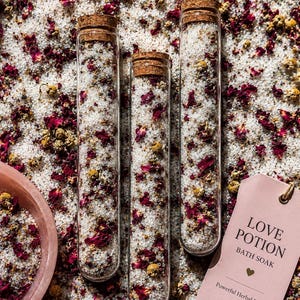 May include: Three clear glass test tubes with cork stoppers filled with white bath salts, dried red rose petals, and yellow chamomile flowers. A light pink tag reads LOVE POTION BATH SOAK Powerful Herbal Blend with a small heart icon.