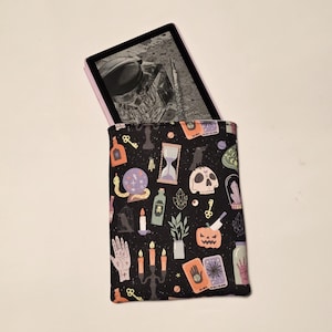 May include: Black e-reader case with a pattern of mystical symbols, including keys, skulls, hourglasses, and potion bottles. The case holds a black and pink e-reader. The overall theme is a witchy or mystical aesthetic.