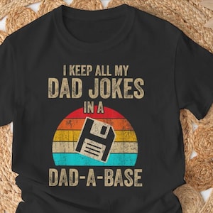 Puede incluir: Camiseta negra con el texto "I KEEP ALL MY DAD JOKES IN A DAD-A-BASE". El diseño presenta un gráfico de disquete de estilo retro sobre un fondo de colores de puesta de sol. El texto y el gráfico son de estilo desgastado.