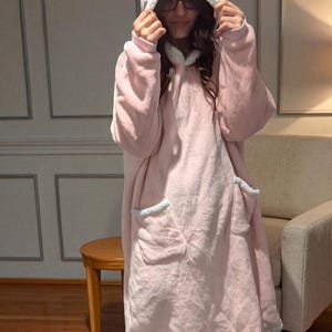 May include: A light pink hooded blanket with a soft, plush texture. The blanket features a hood, large front pockets, and white trim around the hood, pockets, and hem. The person is wearing black socks.
