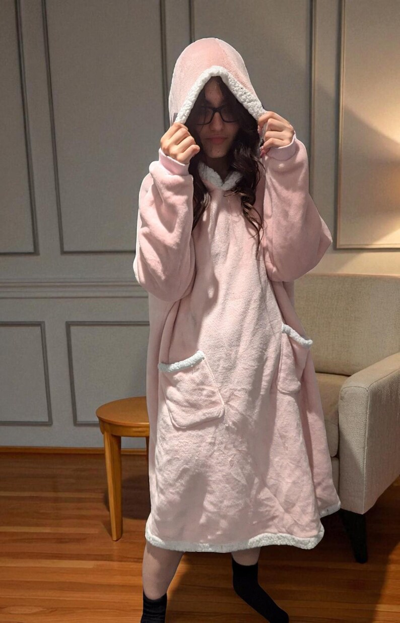 May include: A light pink hooded blanket with a soft, plush texture. The blanket features a hood, large front pockets, and white trim around the hood, pockets, and hem. The person is wearing black socks.