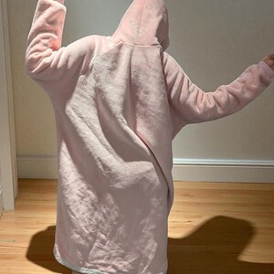 May include: A light pink, hooded blanket with long sleeves. The blanket is made of a soft, plush material and has a white trim around the bottom. The person is wearing the blanket, with their arms raised.