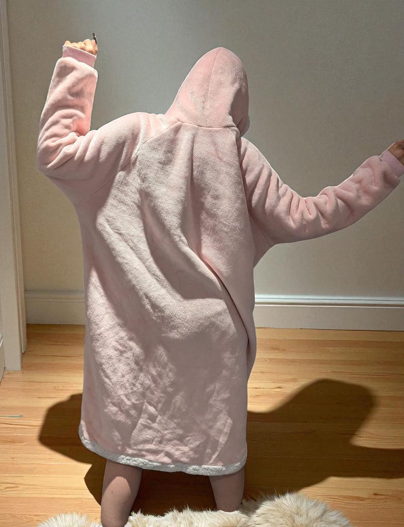 May include: A light pink, hooded blanket with long sleeves. The blanket is made of a soft, plush material and has a white trim around the bottom. The person is wearing the blanket, with their arms raised.