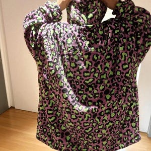 Puede incluir: Una manta con capucha y de gran tama&ntilde;o en un tono p&uacute;rpura con un estampado de leopardo negro y verde. La manta tiene mangas largas y una capucha grande. La persona lleva la manta.