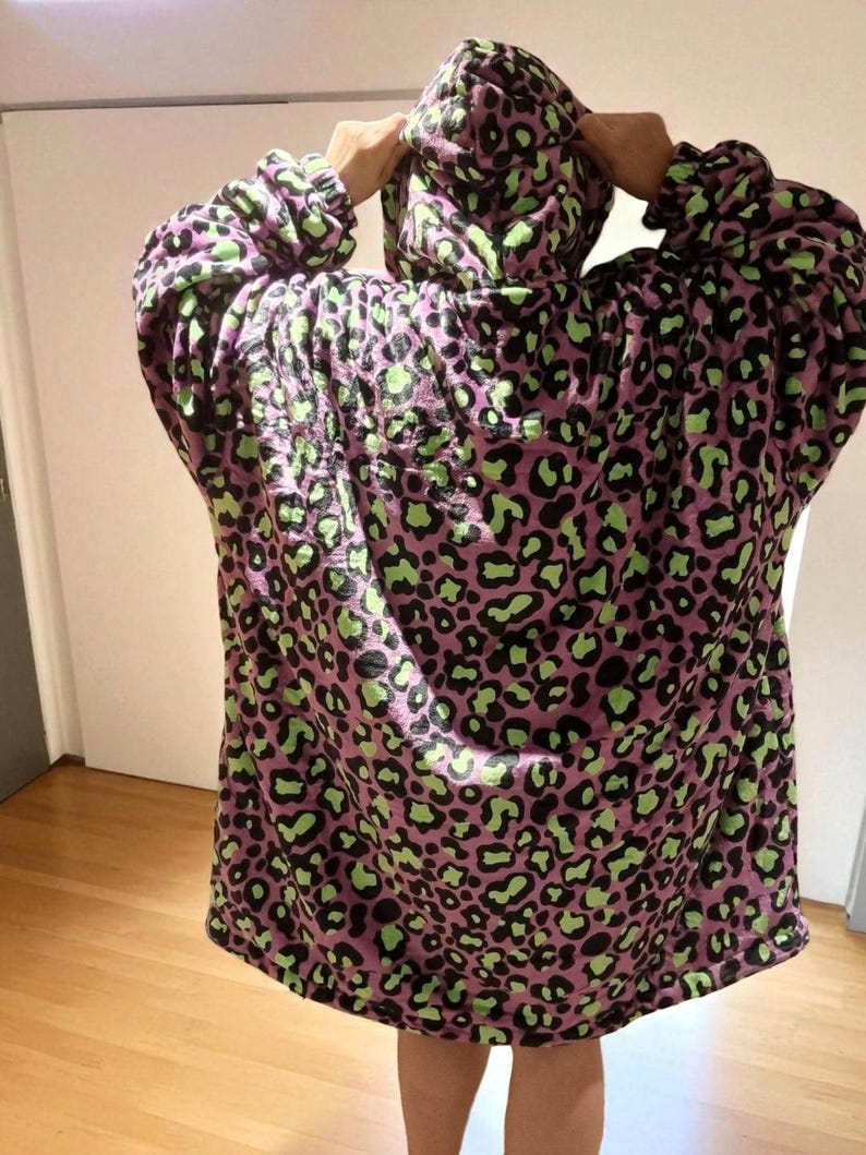 Puede incluir: Una manta con capucha y de gran tama&ntilde;o en un tono p&uacute;rpura con un estampado de leopardo negro y verde. La manta tiene mangas largas y una capucha grande. La persona lleva la manta.