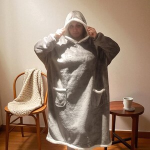 May include: A gray hooded blanket with white trim and two front pockets. The blanket is being worn, and the hood is up. The person is wearing leopard print slippers. A wooden chair with a cream-colored blanket and a small side table are in the background.