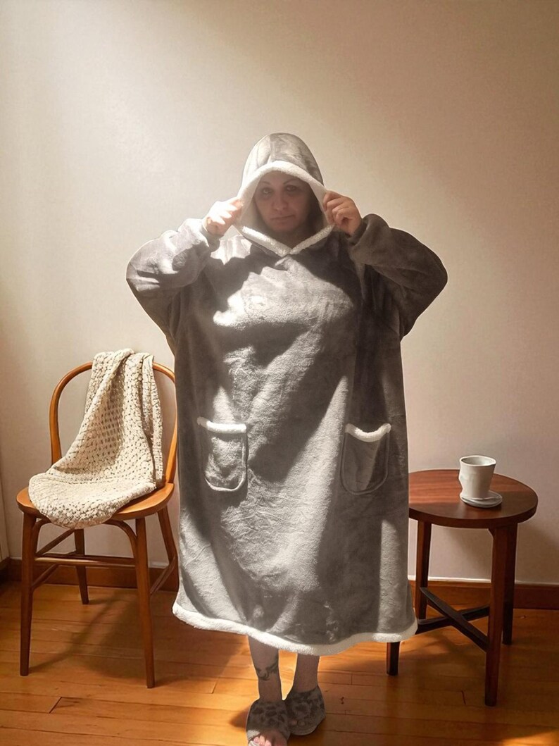 May include: A gray hooded blanket with white trim and two front pockets. The blanket is being worn, and the hood is up. The person is wearing leopard print slippers. A wooden chair with a cream-colored blanket and a small side table are in the background.