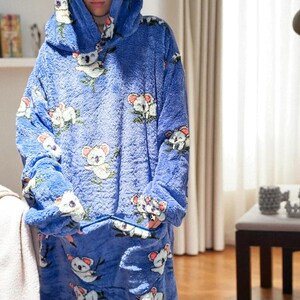 May include: A blue oversized hooded blanket with a koala bear pattern. The blanket features a large hood, long sleeves, and a front pocket. The koala bears are white and gray with pink accents, set against a blue background.