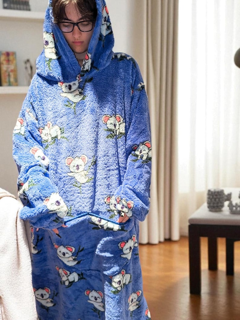 May include: A blue oversized hooded blanket with a koala bear pattern. The blanket features a large hood, long sleeves, and a front pocket. The koala bears are white and gray with pink accents, set against a blue background.