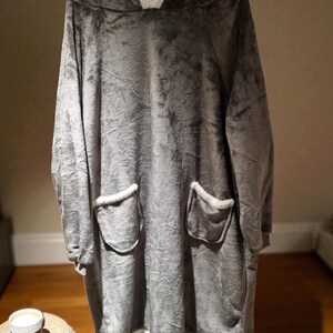 May include: A gray oversized hooded blanket with a plush texture. The blanket features a white fleece-lined hood and pockets, and white trim along the bottom. The cozy wearable blanket is designed for comfort and warmth.