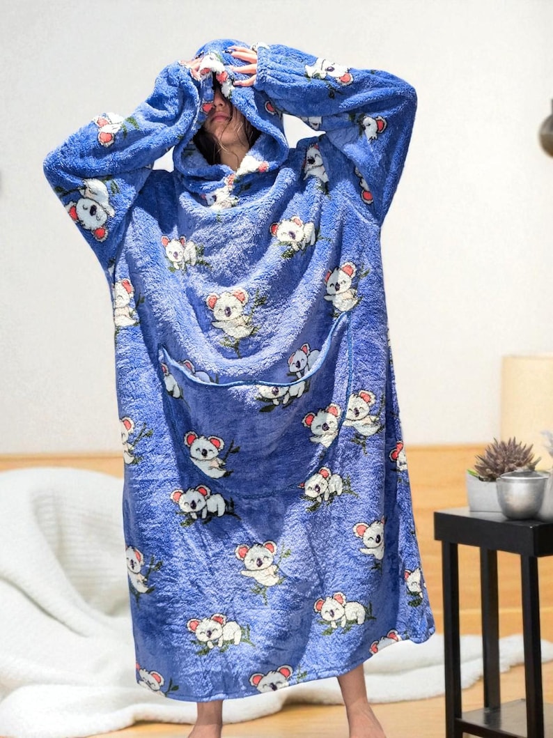 May include: A blue hooded blanket with a koala bear pattern. The blanket has a large front pocket and is made of a soft, plush material. The koalas are white with red accents and are holding green leaves.