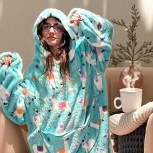 May include: A turquoise hooded blanket with a llama pattern. The oversized wearable blanket features a large front pocket and a soft, plush texture. The person in the image is wearing glasses and is sitting in a chair.