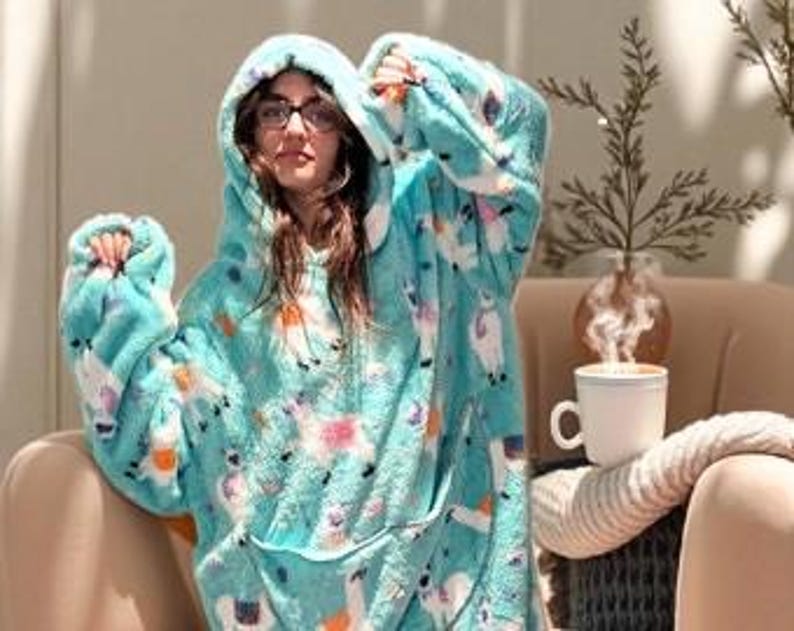May include: A turquoise hooded blanket with a llama pattern. The oversized wearable blanket features a large front pocket and a soft, plush texture. The person in the image is wearing glasses and is sitting in a chair.