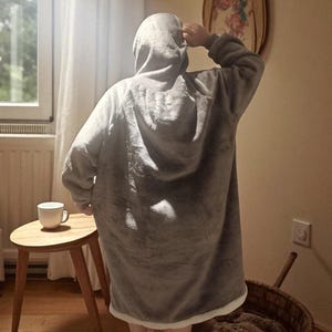 May include: A person wearing a gray hooded blanket, with a white trim, stands in a room. A small wooden side table with a white mug sits nearby. A woven basket and a decorative wall hanging are also visible.