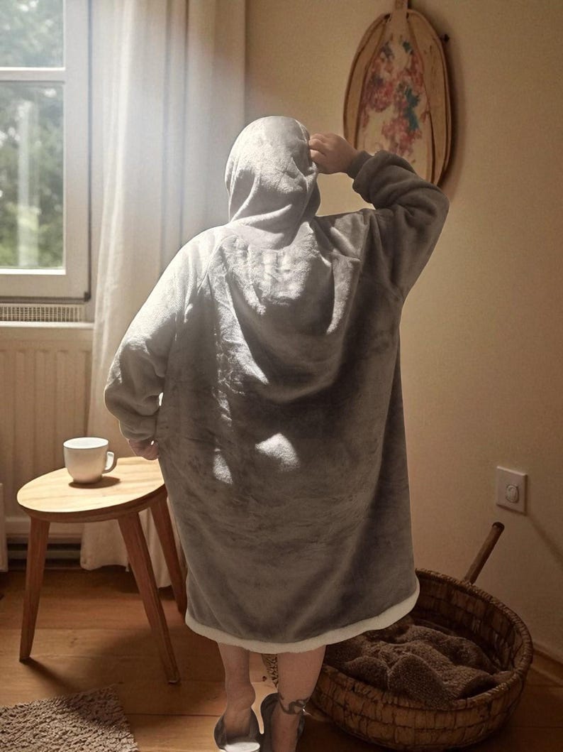 May include: A person wearing a gray hooded blanket, with a white trim, stands in a room. A small wooden side table with a white mug sits nearby. A woven basket and a decorative wall hanging are also visible.