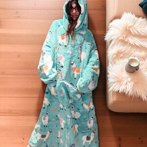 May include: A turquoise hooded blanket with a llama pattern. The blanket has a large front pocket and a hood. The llama pattern includes white, pink, and orange llamas on a turquoise background. The blanket is made of a soft, plush material.