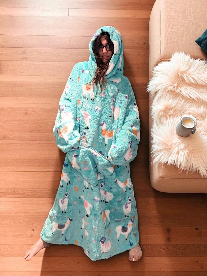 May include: A turquoise hooded blanket with a llama pattern. The blanket has a large front pocket and a hood. The llama pattern includes white, pink, and orange llamas on a turquoise background. The blanket is made of a soft, plush material.