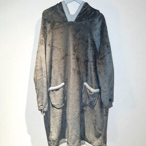 May include: A gray oversized hooded blanket with two front pockets and a hood. The blanket is made of a soft, plush material and has white trim around the pockets and hood. The blanket is hanging on a gray hanger.