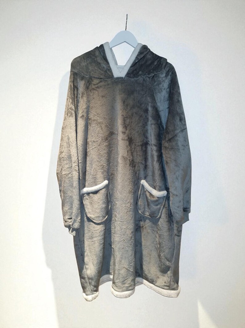 May include: A gray oversized hooded blanket with two front pockets and a hood. The blanket is made of a soft, plush material and has white trim around the pockets and hood. The blanket is hanging on a gray hanger.
