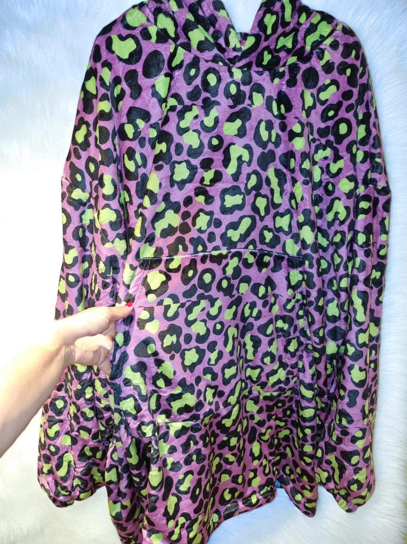 Puede incluir: Una manta con capucha con estampado de leopardo. La manta tiene una base morada con manchas negras y verde ne&oacute;n. Cuenta con una capucha y un gran bolsillo delantero. El material suave y afelpado est&aacute; dise&ntilde;ado para la comodidad y el calor.