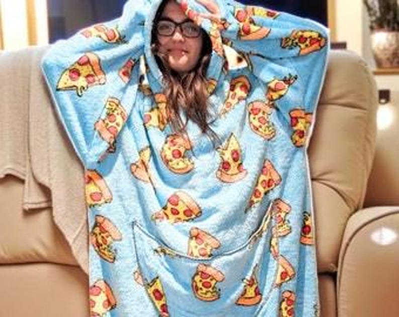 May include: A light blue hooded blanket with a pizza slice pattern. The blanket has a large front pocket and a hood. The pizza slices are yellow, red, and orange. The person wearing the blanket is smiling and wearing glasses.