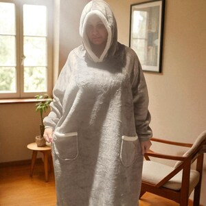 May include: A gray hooded blanket with a white fleece lining and trim. The oversized blanket features a large hood, long sleeves, and two front pockets. The blanket is designed for comfort and warmth.