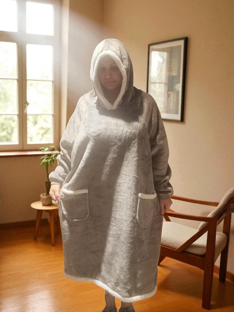 May include: A gray hooded blanket with a white fleece lining and trim. The oversized blanket features a large hood, long sleeves, and two front pockets. The blanket is designed for comfort and warmth.