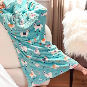 May include: A turquoise fleece blanket with a repeating pattern of white llamas with colorful accents. The blanket is draped over a white couch. The soft blanket has a hood and is designed for comfort and warmth.