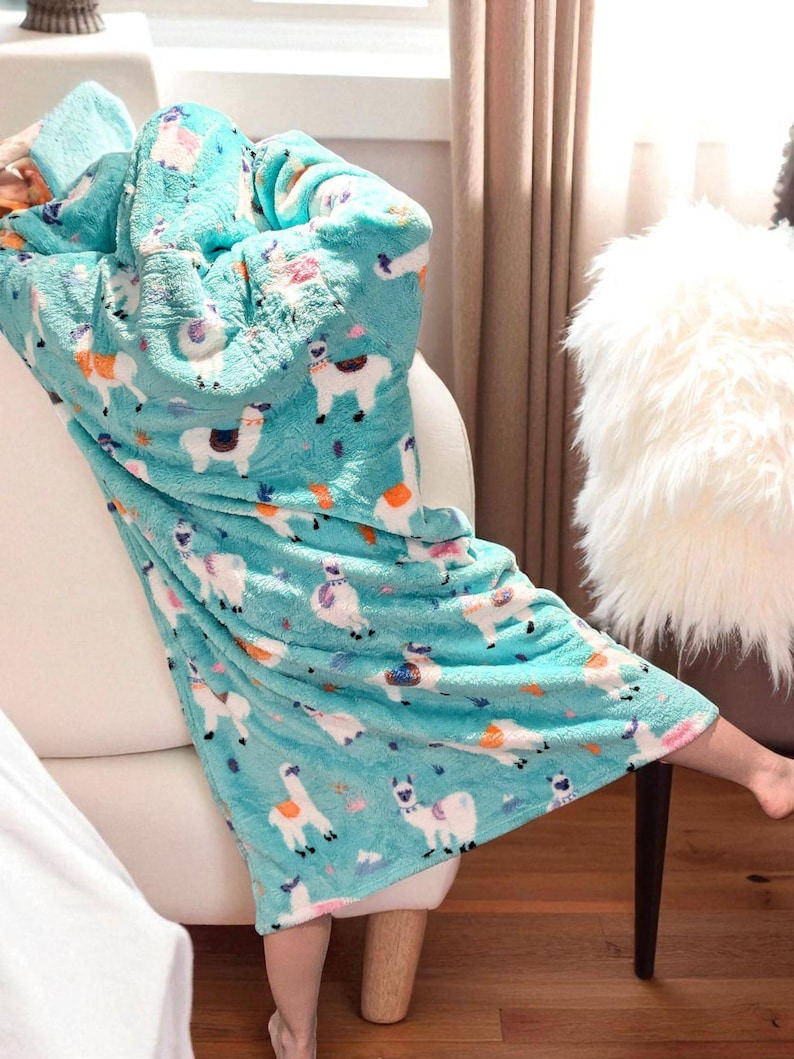 May include: A turquoise fleece blanket with a repeating pattern of white llamas with colorful accents. The blanket is draped over a white couch. The soft blanket has a hood and is designed for comfort and warmth.