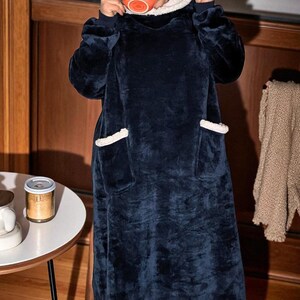 Puede incluir: Una manta con capucha azul marino con forro y ribete de sherpa blanco. La manta tiene dos bolsillos delanteros y se est&aacute; usando. La persona sostiene una taza naranja. La manta est&aacute; dise&ntilde;ada para la comodidad y el calor.