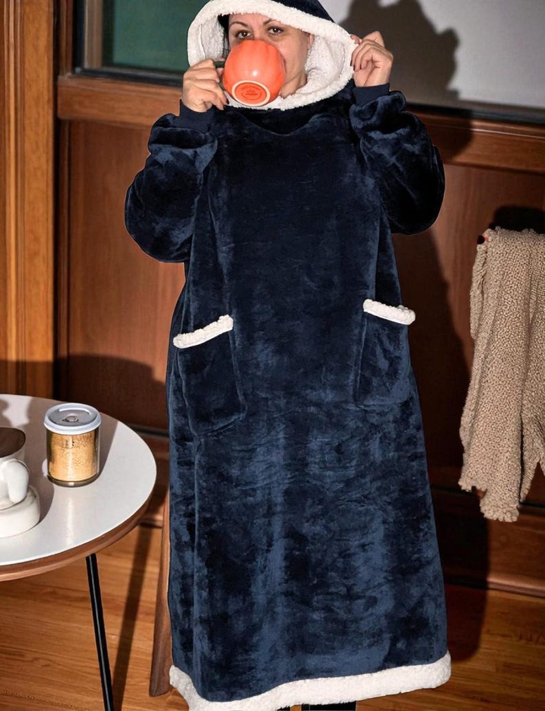 Puede incluir: Una manta con capucha azul marino con forro y ribete de sherpa blanco. La manta tiene dos bolsillos delanteros y se est&aacute; usando. La persona sostiene una taza naranja. La manta est&aacute; dise&ntilde;ada para la comodidad y el calor.