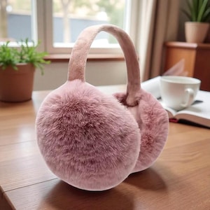May include: Pink faux fur earmuffs with a padded headband. The ear coverings are round and fluffy, designed to keep ears warm. The earmuffs are a soft, muted pink color, suitable for cold weather.