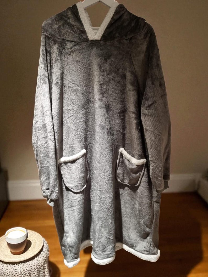 May include: A gray oversized hooded blanket with a plush texture. The blanket features a white fleece-lined hood and pockets, and white trim along the bottom. The blanket is designed for comfort and warmth.