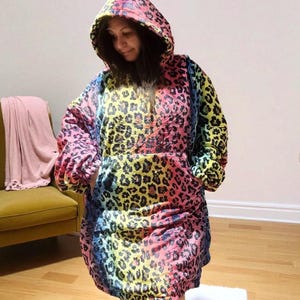 Puede incluir: Una manta con capucha y de gran tama&ntilde;o con estampado de leopardo arco&iacute;ris. La manta tiene una capucha grande, mangas largas y un bolsillo de canguro. La combinaci&oacute;n de colores incluye rosa, amarillo, azul y verde, con manchas de leopardo negras.