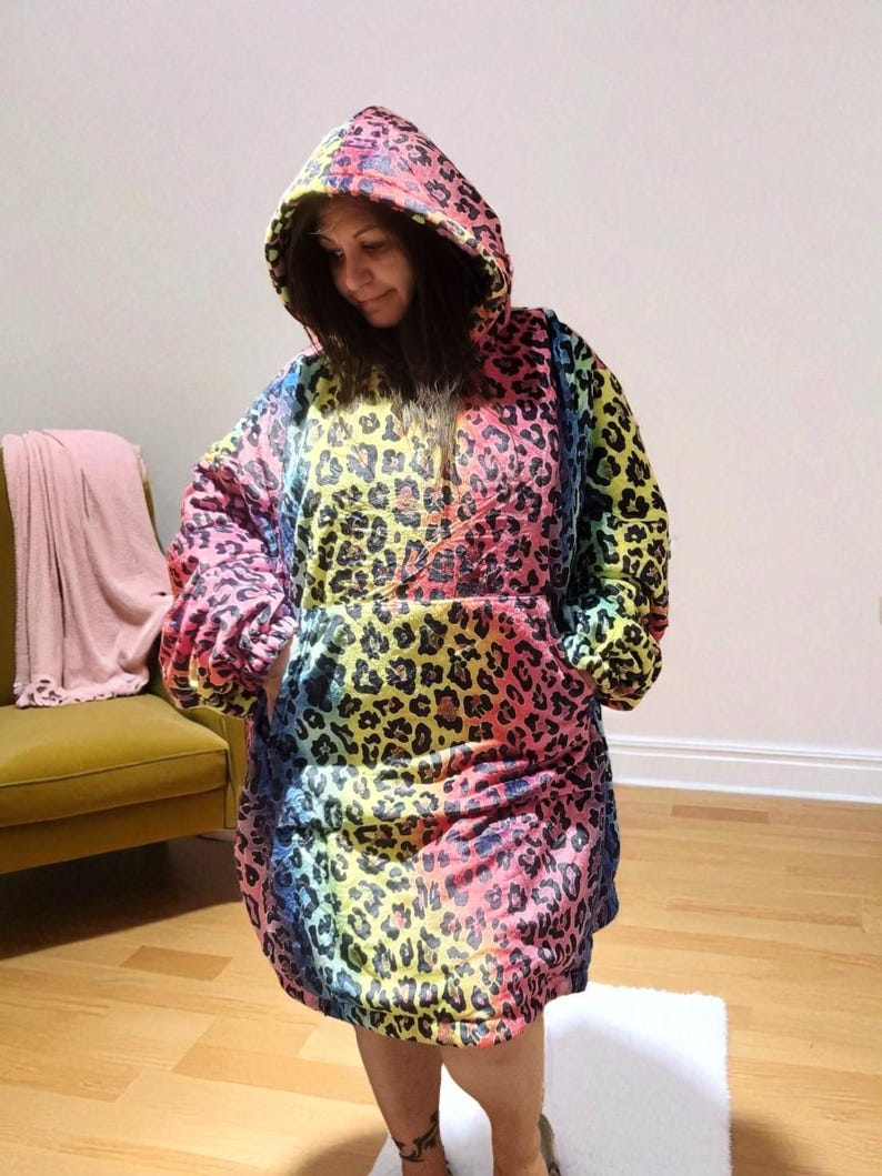 Puede incluir: Una manta con capucha y de gran tama&ntilde;o con estampado de leopardo arco&iacute;ris. La manta tiene una capucha grande, mangas largas y un bolsillo de canguro. La combinaci&oacute;n de colores incluye rosa, amarillo, azul y verde, con manchas de leopardo negras.