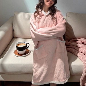 May include: A light pink hooded blanket with white trim and pockets. A black and white coffee cup sits on a small brown saucer. A pink knitted blanket is draped on the sofa.