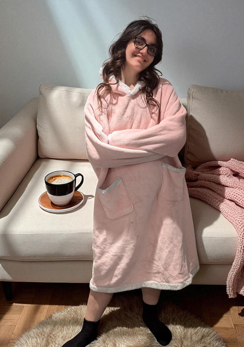 May include: A light pink hooded blanket with white trim and pockets. A black and white coffee cup sits on a small brown saucer. A pink knitted blanket is draped on the sofa.