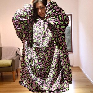 Puede incluir: Una manta con capucha y de gran tama&ntilde;o con estampado de leopardo. La manta presenta una base de color lila claro con manchas negras y verde ne&oacute;n. Tiene un gran bolsillo frontal y mangas largas, ofreciendo un aspecto acogedor.