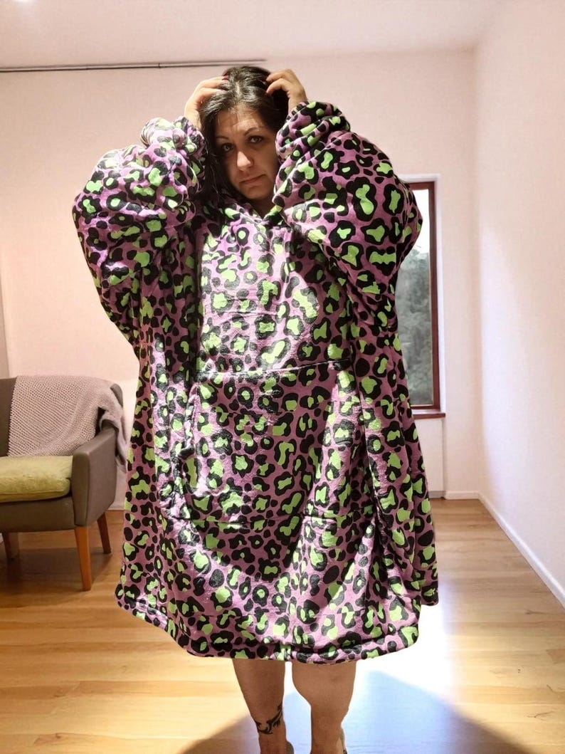 Puede incluir: Una manta con capucha y de gran tama&ntilde;o con estampado de leopardo. La manta presenta una base de color lila claro con manchas negras y verde ne&oacute;n. Tiene un gran bolsillo frontal y mangas largas, ofreciendo un aspecto acogedor.