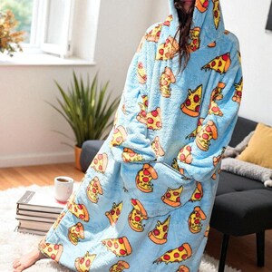May include: A light blue, hooded blanket with a pizza slice pattern. The blanket features a large front pocket and a hood. The pizza slices have yellow crusts, red pepperoni, and orange cheese. The blanket is worn by a person.