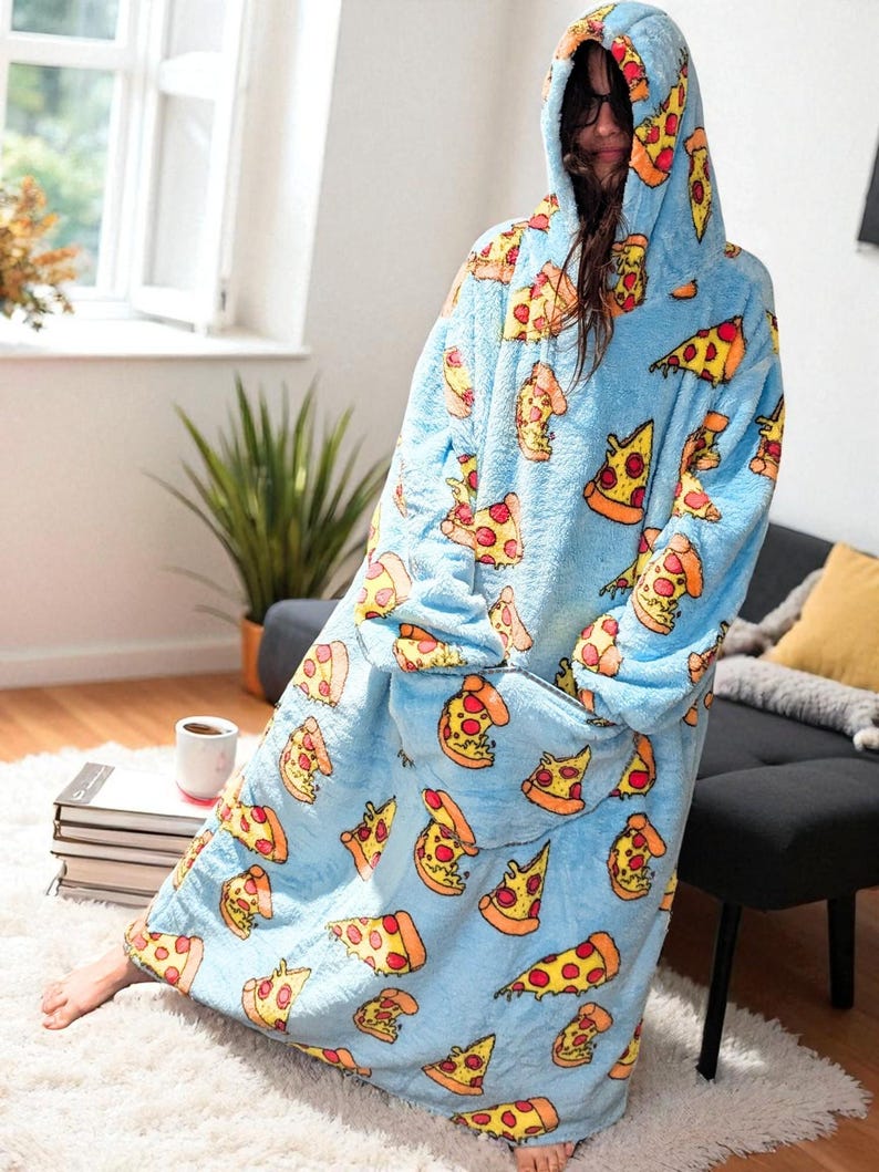 May include: A light blue, hooded blanket with a pizza slice pattern. The blanket features a large front pocket and a hood. The pizza slices have yellow crusts, red pepperoni, and orange cheese. The blanket is worn by a person.